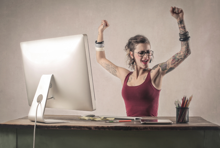 A happy woman with extensive tattoos sits at a desk in from of a computer screen and raises her arms in celebration