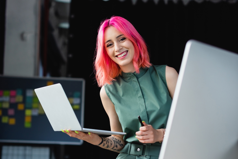 A smiling girl with pink and orange hair, neon yellow nails, and a tattoo sleeve holds a laptop in her hand and smiles at the camera