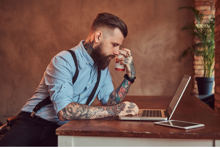 Young man with bright tattoos on both arms holds a cup of coffee and works on a laptop, with a tablet nearby