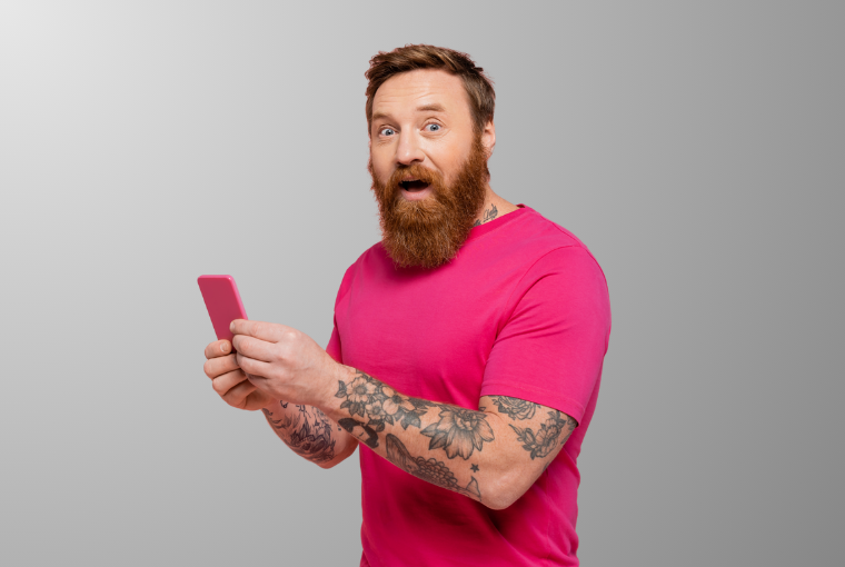 Surprised man in a bright pink T-shirt and tattoos on his arm looks at the camera holding a bright pink phone in both hands