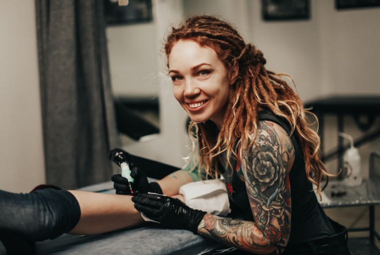 A confident female tattoo artist smiles at the camera mid-design.