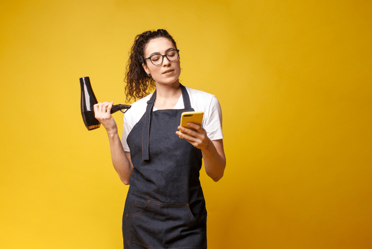 A hairdresser stands against a bright yellow background with a hair dryer in one had and a mobile phone in the other