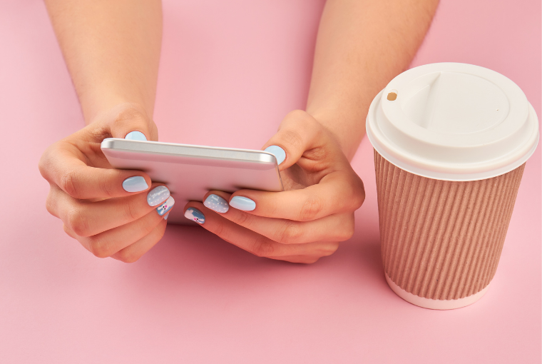Two hands with blue manicured nails hold a phone next to a takeaway coffee cup