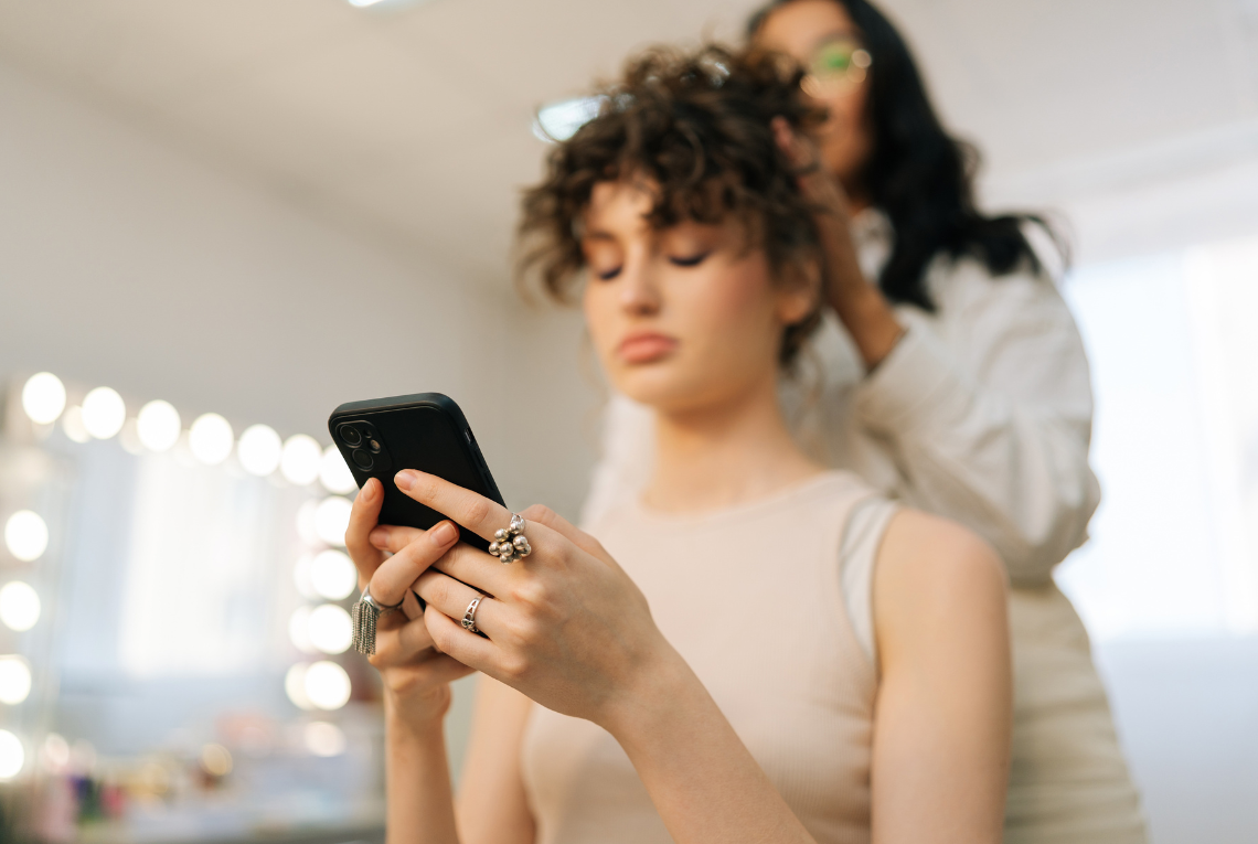 Young woman scrolls on her phone while a hairdresser styles her hair at the salon