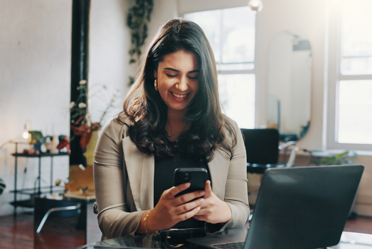 A smiling salon owner sits in front of a laptop as she sets up her salon website on her mobile phone