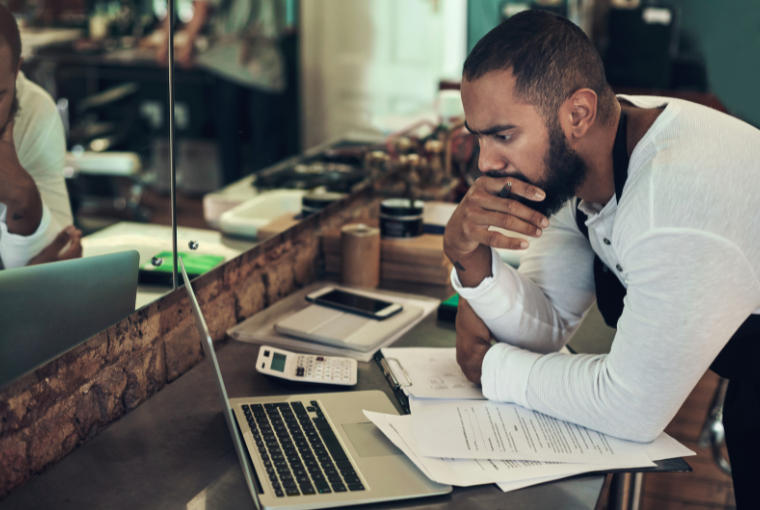 A male hair stylist frowns as he leans over his laptop. he also has a pile of papers and a calculator to hand