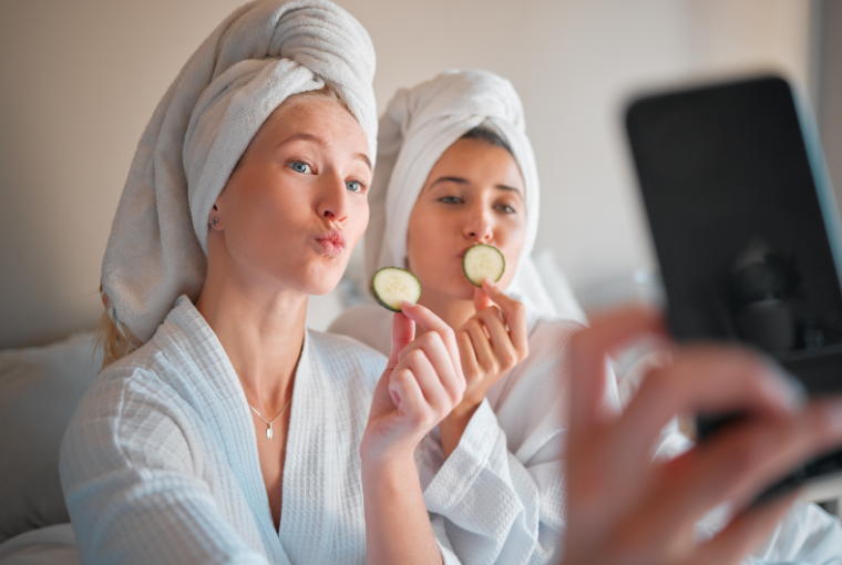 Two young women in white robes and towels on their heads hold a slice of cucumber as they pose for a fun selfie at the spa